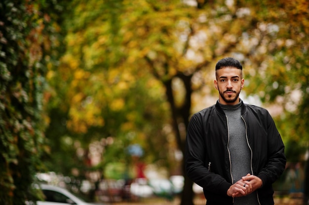 Il ritratto di un uomo elegante con barba araba indossa un dolcevita grigio e una giacca nera Ragazzo modello arabo su uno sfondo di foglie autunnali