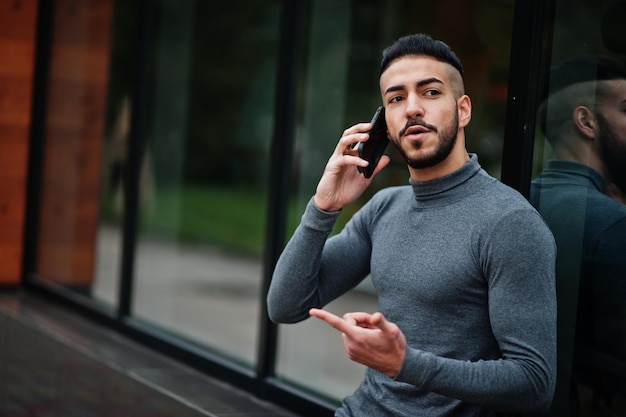 Il ritratto di un uomo elegante con barba araba indossa un modello arabo a collo alto grigio seduto e parla con lo smartphone