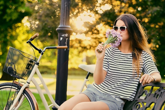 Il ritratto di una bruna attraente si siede su una panchina con una bicicletta in un parco cittadino.