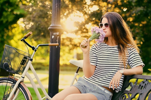 Il ritratto di una bruna attraente si siede su una panchina con una bicicletta in un parco cittadino.