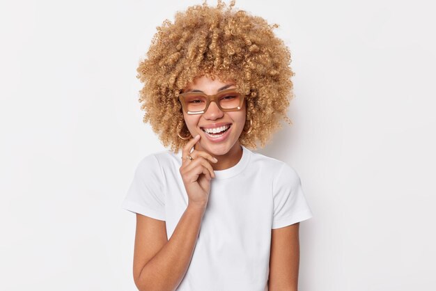 Il ritratto di una donna dai capelli ricci allegra sorride ampiamente mantiene il dito vicino alla bocca essendo di buon umore esprime emozioni positive indossa occhiali e maglietta casual isolata su sfondo bianco