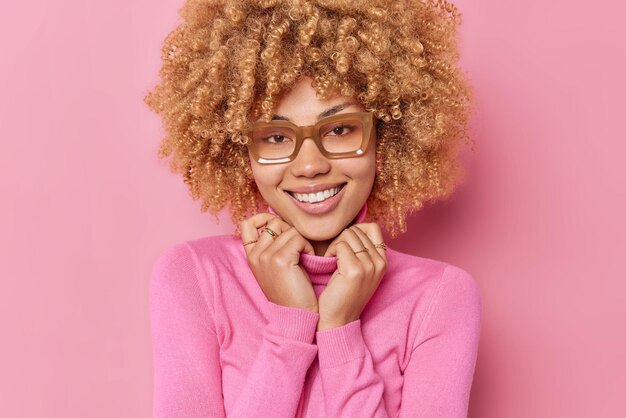 Il ritratto di una donna dai capelli ricci sorride delicatamente tiene le mani sul colletto del maglione esprime emozioni positive sente buone notizie indossa occhiali isolati su sfondo rosa. Concetto di emozioni felici