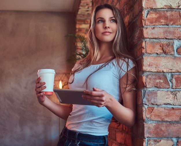 Il ritratto di una giovane donna bionda tiene una tazza di caffè da asporto e una tavoletta digitale mentre si appoggia a un muro di mattoni in una stanza con interni soppalcati.