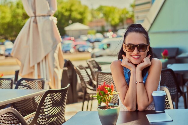 Il ritratto di una ragazza bruna affascinante e felice che indossa abiti alla moda si sta godendo la giornata estiva mentre è seduto su una terrazza in un caffè all'aperto, guardando la telecamera.