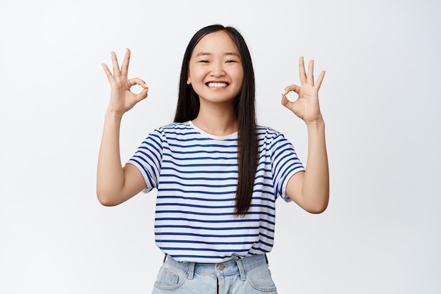 Il ritratto di una ragazza bruna asiatica mostra segni ok e sorride soddisfatto, feedback positivo, piace e approva smth buono, sta su bianco.