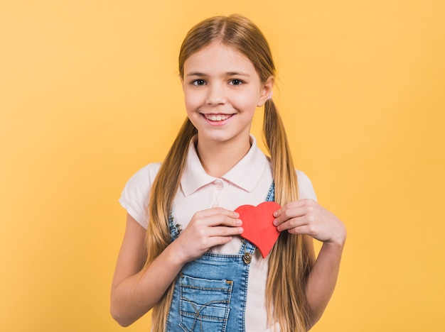 Il ritratto di una ragazza sorridente con capelli lunghi biondi che mostrano la carta rossa ha tagliato il cuore che sta contro il fondo giallo