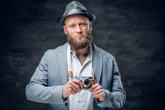 Il ritratto in studio del fotografo barbuto vestito con un abito e un cappello di feltro tiene una vecchia macchina fotografica SLR vintage.