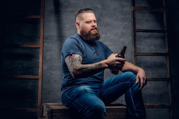 Il ritratto in studio del maschio hipster tatuato barbuto tiene la bottiglia di birra su sfondo grigio con due scale di legno vintage.