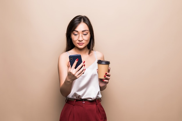 Il ritratto integrale della donna asiatica sorridente che per mezzo del telefono cellulare mentre tiene la tazza di caffè per andare ha isolato sopra la parete beige