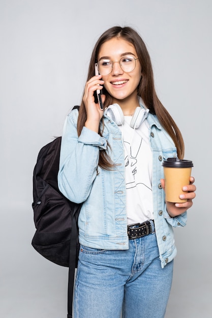 Il ritratto laterale di giovane donna dello studente parla con smartphone, tenendo il caffè per andare tazza isolata sulla parete grigia