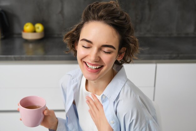 Il ritratto mattutino felice di una donna castana felice beve una tazza di caffè nella sua cucina e sorride accogliente