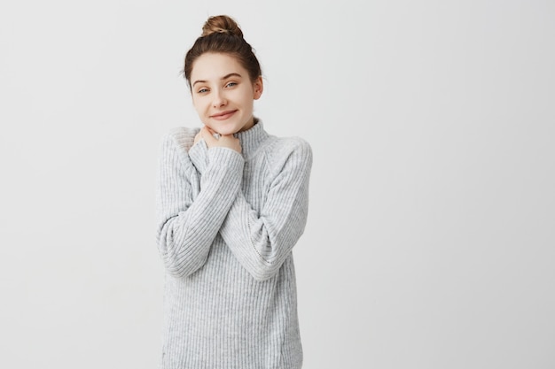 Il ritratto orizzontale della donna adorabile ha legato i suoi capelli nel topknot che sorride essendo positivo. Bello parrucchiere femminile che ha rottura che mette le mani sul petto nella gioia. Tempo libero, concetto di persona