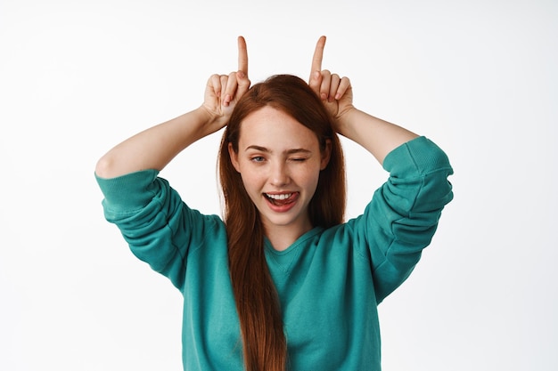 Il ritratto ravvicinato della ragazza dai capelli rossi impertinente mostra il gesto delle corna di toro, gli strizzatine d'occhio e i sorrisi audaci, dall'aspetto determinato e sicuro di sé, divertendosi, sfondo bianco