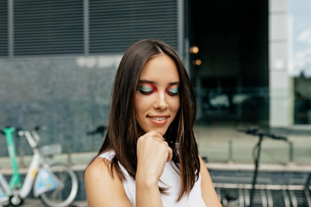 Il ritratto ravvicinato di una bella donna alla moda con un trucco luminoso per le vacanze sta guardando in basso e sorride all'aperto alla luce del sole