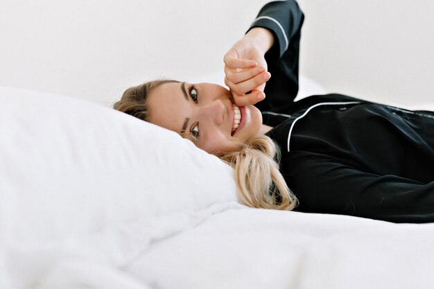 Il ritratto ravvicinato di una donna adorabile e romantica in abiti da casa sorride ampiamente e si tocca le labbra mentre posa davanti alla telecamera nel letto