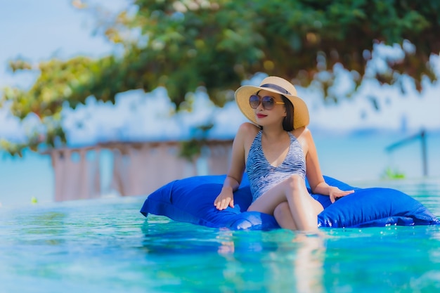 Il sorriso felice della bella giovane donna asiatica del ritratto si rilassa nella piscina alla spiaggia vicina dell&#39;oceano del mare della località di soggiorno dell&#39;hotel sul cielo blu