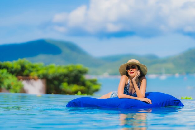 Il sorriso felice della bella giovane donna asiatica del ritratto si rilassa nella piscina alla spiaggia vicina dell&#39;oceano del mare della località di soggiorno dell&#39;hotel sul cielo blu