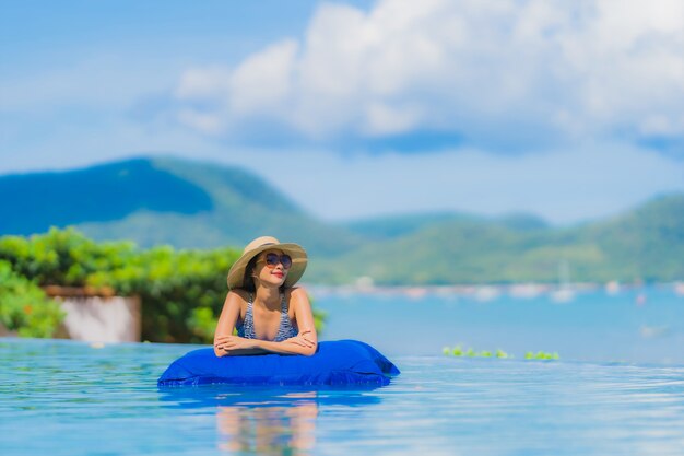 Il sorriso felice della bella giovane donna asiatica del ritratto si rilassa nella piscina alla spiaggia vicina dell&#39;oceano del mare della località di soggiorno dell&#39;hotel sul cielo blu