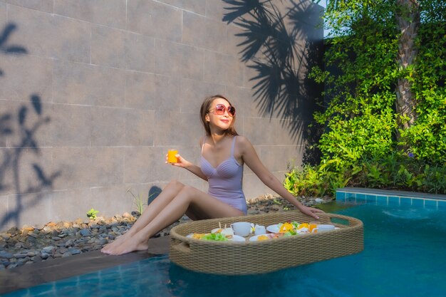 Il sorriso felice della giovane donna asiatica del ritratto gode di con il vassoio di galleggiamento della prima colazione nella piscina in hotel