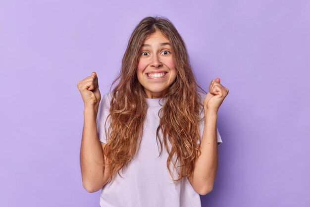 Il tiro al coperto di una giovane donna europea felice stringe i pugni e morde le labbra in attesa di importanti risultati positivi vestita con una maglietta casual isolata sul muro viola