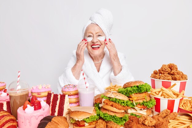 Il tiro al coperto di una vecchia signora allegra applica cerotti sotto gli occhi per ridurre le rughe ha sorrisi di manicure rossi ampiamente di buon umore mangia grandi porzioni di fast food