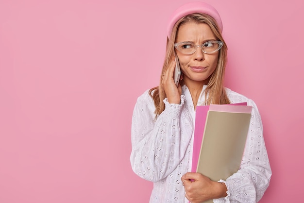 Il tiro orizzontale di una giovane studentessa europea dispiaciuta tiene libri di testo e fa una telefonata ha un'espressione del viso infelice indossa occhiali e camicetta bianca isolata sul rosa