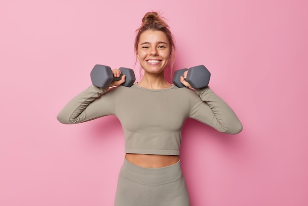 Il tiro orizzontale di una sportiva motivata soddisfatta ha un allenamento regolare alza le braccia con i manubri fa esercizi per i muscoli vestiti con abbigliamento sportivo sorride isolato sul muro rosa. Concetto di sport