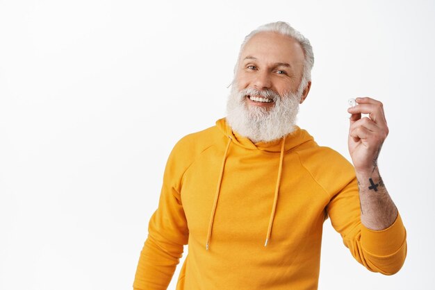 Il vecchio hipster sorridente si toglie le cuffie e sembra felice metti un auricolare nell'orecchio per ascoltare la musica e fare una telefonata utilizzando auricolari wireless in piedi su sfondo bianco