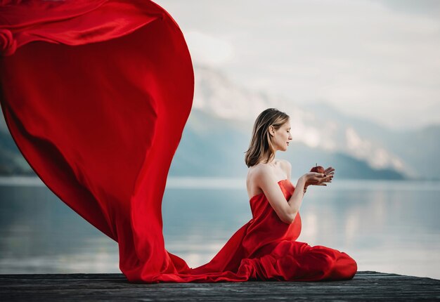 Il vento soffia il vestito rosso di una donna incinta che si siede con la mela sul ponte sul lago