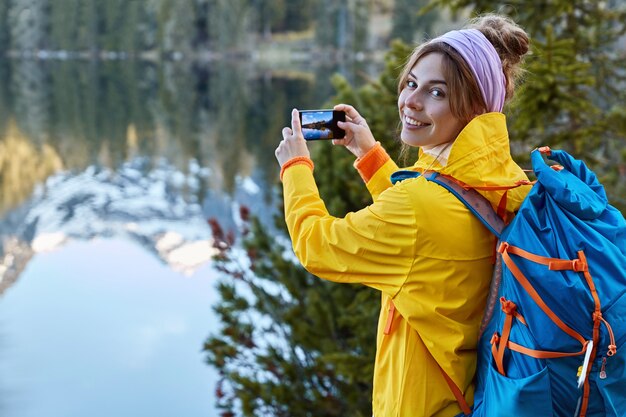 Il viaggiatore turistico tiene in mano lo smart phone, fa foto del paesaggio panoramico in viaggio, ammira il viaggio in montagna, posa vicino al lago