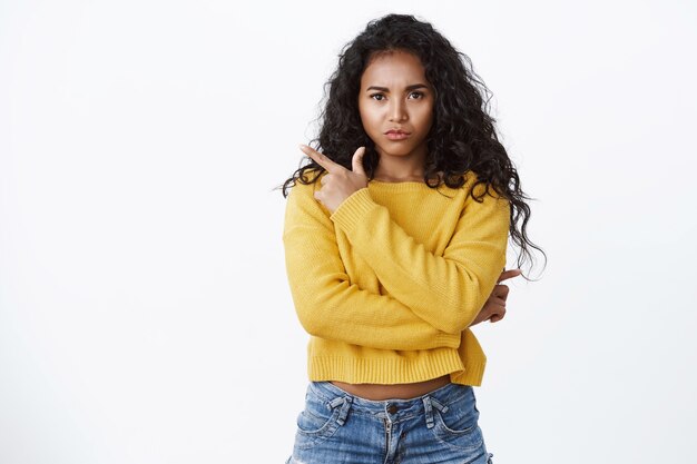 Imbronciata, delusa ragazza afroamericana con i capelli ricci, indossa jeans, maglione giallo, sopracciglia aggrottate e labbra della borsa offese, indicando l'angolo in alto a sinistra sconvolto e insoddisfatto