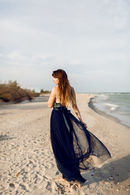 Immagine a tutta altezza della donna alla moda in abito di lusso elegante in posa sulla spiaggia. Vista dal retro. Peli lunghi.