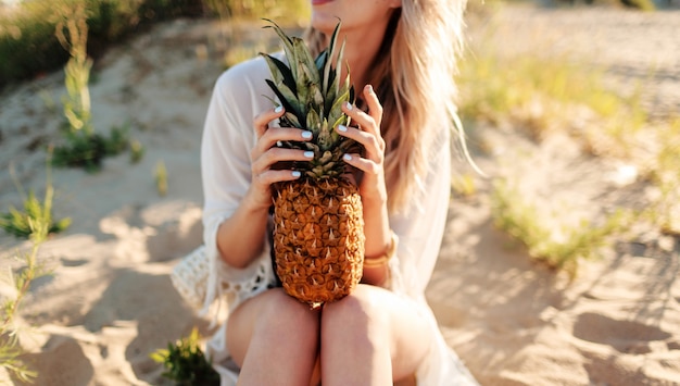 Immagine all'aperto di stile di vita di ridere bella donna con ananas succoso rilassante sulla spiaggia assolata. Vestito estivo alla moda