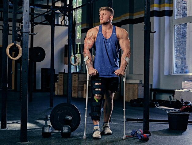 Immagine del corpo intero di un enorme bodybuilder con le stampelle in una palestra.