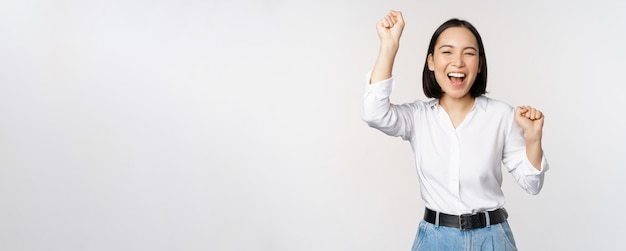 Immagine del gesto felice e fortunato della donna asiatica evviva vincendo e celebrando trionfante alzando le mani e ridendo in piedi su sfondo bianco