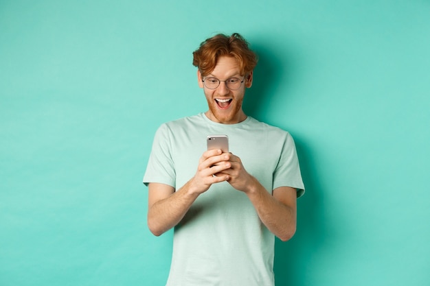 Immagine del giovane uomo dai capelli rossi con gli occhiali che legge lo schermo del telefono con la faccia sorpresa, riceve un'offerta promozionale incredibile, in piedi su sfondo turchese