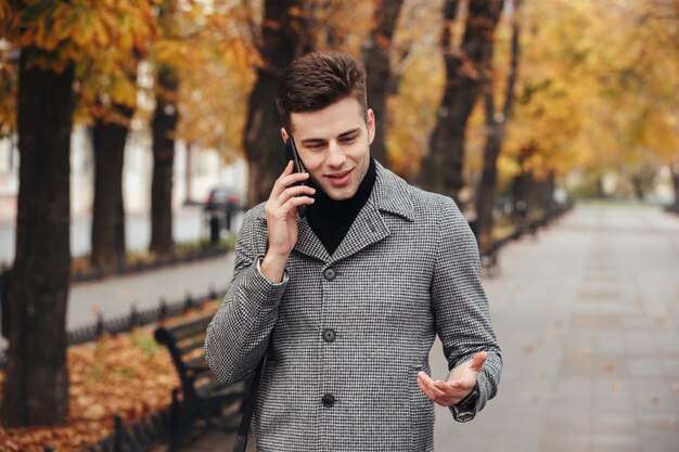 Immagine del maschio elegante in cappotto che cammina nel parco vuoto con gli alberi di autunno e che parla sullo smartphone