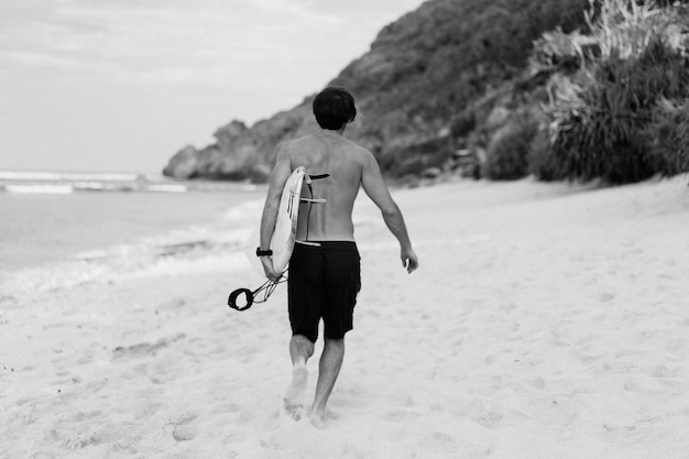 Immagine del paesaggio del surfista maschio impegnato a camminare sulla spiaggia all'alba mentre trasportava la sua tavola da surf sotto il braccio con le onde dell'oceano che si infrangono sullo sfondo. Giovane surfista maschio bello sull'oceano