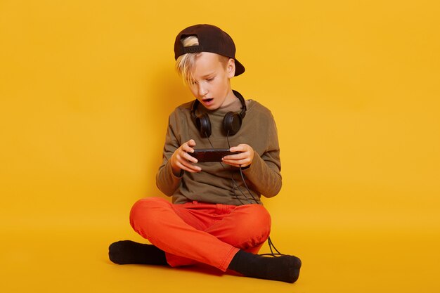 Immagine del ragazzo che gioca sul telefono cellulare. Scherzi la seduta sul pavimento in studio isolato sopra sulla parete gialla e tenendo il telefono cellulare in mano, giocando il suo gioco online preferito, tiene le gambe incrociate.