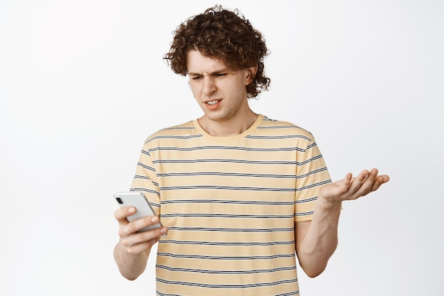 Immagine del ragazzo riccio che fissa confuso il telefono cellulare in piedi in maglietta su sfondo bianco