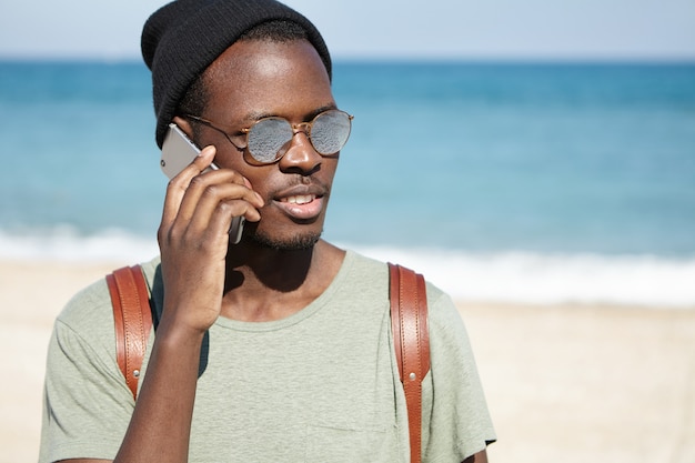 Immagine del turista maschio nero sembrante d'avanguardia con lo zaino, il cappello d'uso e gli occhiali da sole su tempo soleggiato, parlando sul telefono cellulare