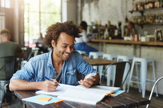 Immagine dell'elegante studente africano con l'orecchino che indossa la camicia di jeans seduto al tavolo di legno a fare i compiti tenendo lo smartphone felice di ricevere un messaggio dal suo amico digitando qualcosa