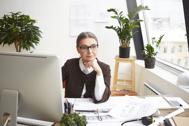 Immagine dell'ingegnere costruttore femminile maturo esperto premuroso in occhiali alla moda con sguardo pensieroso durante lo sviluppo della documentazione del progetto di costruzione, seduto alla scrivania davanti al computer