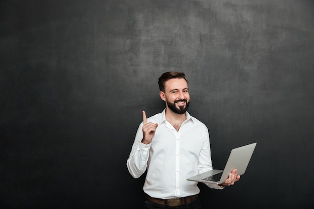 Immagine dell'uomo castana astuto che lavora nell'ufficio facendo uso del computer portatile d'argento che gesturing con il dito su, isolato sopra la parete grigio scuro