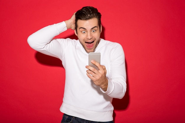 Immagine dell'uomo felice sorpreso in maglione che esamina smartphone sopra la parete rossa