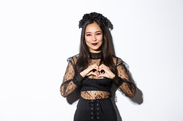 Immagine della bella donna asiatica sorridente che celebra Halloween, mostrando il gesto del cuore e guardando la telecamera, in piedi su sfondo bianco in abito di pizzo gotico con corona.