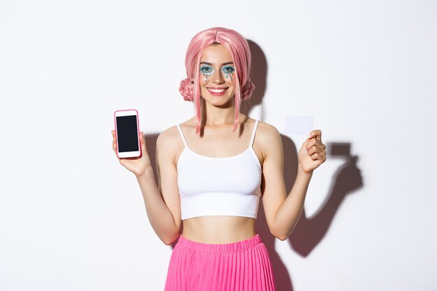 Immagine della bella ragazza sorridente in parrucca rosa, con trucco luminoso, che mostra la carta di credito e lo schermo del telefono cellulare, in piedi.