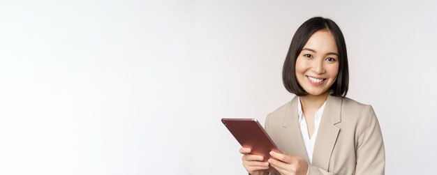 Immagine della commessa asiatica della donna di affari che tiene la compressa digitale e che sorride lavorando con il supporto del gadget