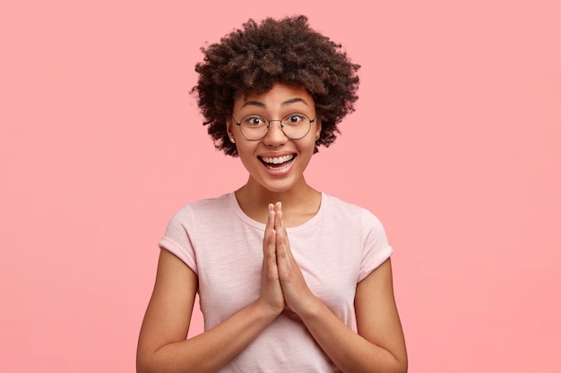 Immagine della donna afroamericana felice con un sorriso piacevole, vestita casualmente, preme le mani insieme, posa in gesto di preghiera, implora perdono con espressione gioiosa, isolato sul muro rosa