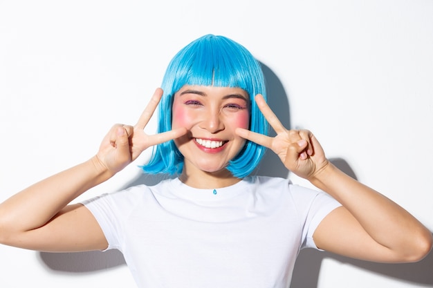 Immagine della donna asiatica carina in parrucca blu che mostra gesto di pace kawaii e sorridente